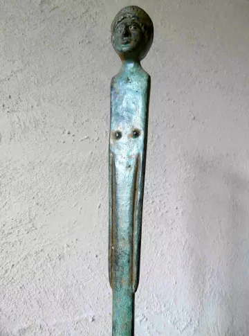 L'image montre un objet en métal, probablement en bronze, qui semble être une sorte de sculpture ou de barre. La partie supérieure représente une tête humaine stylisée, tandis que la tige qui descend présente des éléments qui pourraient évoquer un corps. La surface a une patine verdâtre, indiquant son ancienneté. L'ensemble de l'objet évoque une forme artistique, fusionnant des éléments figuratifs et abstraits.