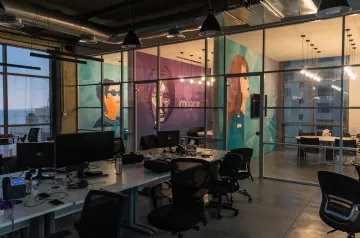The image shows a modern office space with large glass windows, providing a view of the outside. Inside, there are several workstations with computers and ergonomic chairs. The walls are decorated with vibrant murals featuring stylized portraits of individuals, adding a creative touch to the environment. The overall ambiance is open and contemporary, with industrial elements like exposed ducts and warm lighting fixtures. The space appears to be designed for collaboration and innovation.