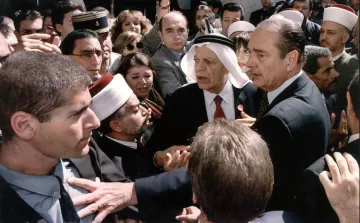 L'image montre une scène chargée d'émotion et de tension. On y voit plusieurs personnes rassemblées autour d'une figure centrale, visiblement un homme en costume. D'autres personnes, portant des vêtements traditionnels et des turbans, semblent interagir avec lui. L'atmosphère est dynamique, avec des visages concentrés et des gestes animés, ce qui suggère une discussion ou un événement important. Le mélange de styles vestimentaires indique une diversité culturelle parmi les personnes présentes, ce qui pourrait refléter un moment significatif sur le plan social ou politique.