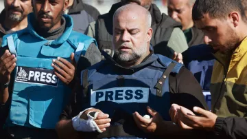 En la imagen, un grupo de personas está reunido, y se puede notar a un hombre en el centro que lleva un chaleco con la palabra "PRESS" (prensa) en grandes letras. Este hombre tiene una expresión de concentración y parece estar rezando. Su mano derecha está vendada. Los demás en el grupo también tienen una actitud similar, algunos con las manos levantadas en señal de oración. El ambiente sugiere un momento de solemnidad o de conmemoración.