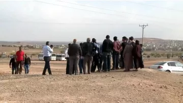 L'image montre un groupe de personnes rassemblées sur un terrain semi-aride. Ils semblent observer quelque chose au loin, peut-être un événement ou une scène particulière. L'arrière-plan est dégagé, avec des collines et des bâtiments visibles au loin. Il y a des véhicules stationnés, dont une voiture à droite. L'atmosphère suggère une interaction entre les gens, qui semblent concentrés sur ce qu'ils regardent. Des câbles électriques sont visibles, ajoutant à l'ambiance rurale de la scène.