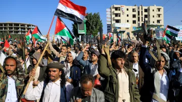 تظهر الصورة مجموعة من الأشخاص في تظاهرة حاشدة، يحملون الأعلام ويرتدون زيًّا تقليديًا. تعبر الوجوه عن الحماس وتظهر الأعلام التي تحمل ألوان الوطن. تتواجد خلفهم بنايات وبعض العلامات التجارية، مما يوحي بأن المكان هو منطقة حضرية. يبدو أن المتظاهرين يمارسون حقهم في التعبير عن آرائهم والمشاركة في حدث سياسي أو اجتماعي.
