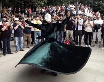 L'image montre une scène de rassemblement en plein air où une personne vêtue d'une longue robe vert foncé tourne gracieusement. Cette personne porte un masque à gaz, ce qui ajoute une dimension étrange et intrigante à la performance. Autour, une foule observe, certains prenant des photos avec leurs téléphones. L'ambiance semble à la fois festive et solennelle, avec des expressions variées parmi les spectateurs. Les arbres en arrière-plan ajoutent une touche de nature à la scène.