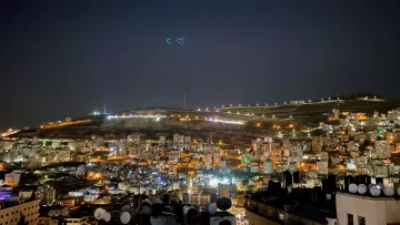 L'image montre une vue nocturne d'une ville illuminée. On peut voir de nombreux bâtiments agencés en terrasses, avec des lumières qui scintillent dans l'obscurité. À l'arrière-plan, des collines sont éclairées, et quelques structures sont visibles au sommet. L'atmosphère est tranquille, avec un ciel nocturne clair. Les antennes et structures en premier plan ajoutent une dimension urbaine à cette scène paisible.