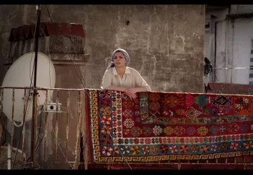 L'image montre une femme se tenant sur un balcon, regardant au loin. Elle porte un haut clair et un foulard sur la tête. Derrière elle, un grand tapis coloré est accrochée à la balustrade, avec des motifs détaillés et vibrants. En arrière-plan, on aperçoit un mur et une antenne satellite, ce qui suggère un environnement urbain. L'éclairage donne une atmosphère chaleureuse à la scène.
