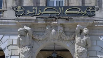 The image features a historical bank building with an ornate entrance. Above the doorway, there is a sign in Arabic that reads "بنك الجزائر" which translates to "Bank of Algeria." The architecture includes decorative elements, such as sculptures of two male figures on either side of the entrance. These figures appear to be holding or surrounded by garlands or fruit, adding to the classical decorative style of the building. The overall design is elaborate, reflecting a historical architectural style.