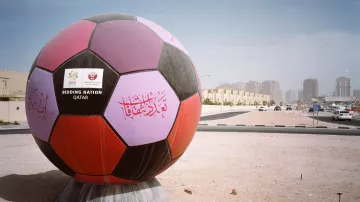 L'image montre un énorme ballon de football décoré, affichant des inscriptions colorées en arabe et d'autres éléments graphiques. Ce ballon est installé sur un terrain, probablement dans un contexte urbain, avec des bâtiments en arrière-plan. Le ciel est clair, et l'environnement semble être une zone en développement ou un lieu préparé pour un événement, suggérant une connexion avec le football ou un événement sportif marquant, tel qu'une compétition internationale.