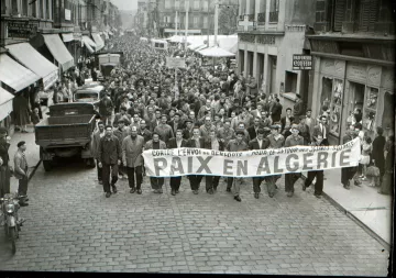 L'image montre une grande foule rassemblée dans une rue, probablement lors d'une manifestation. Les personnes marchent en portant une grande banderole qui indique "PAIX EN ALGÉRIE". On peut voir des hommes et quelques femmes, habillés de manière vestimentaire typique de l'époque. Des voitures et des commerces sont visibles en arrière-plan, ce qui donne une idée du contexte urbain. L'atmosphère semble sérieuse, suggérant que la manifestation est organisée autour d'un sujet important, en l'occurrence la paix en Algérie.