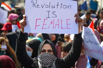 الصورة تظهر مجموعة من المتظاهرين في تجمع حاشد، حيث تحمل امرأة لافتة مكتوب عليها "الثورة أنثوية". يتواجد أفراد من مختلف الأعمار في الخلفية، كما يظهر بعضهم يرتدي الأوشحة والقبعات. الأجواء تعكس حماس المتظاهرين والتزامهم بقضية معينة.