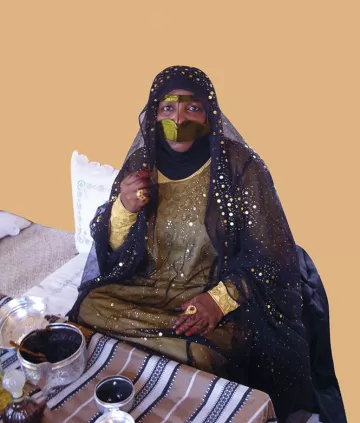 L'image montre une femme vêtue d'une tenue traditionnelle ornée de motifs et de broderies. Elle porte un voile noir qui couvre son visage en grande partie, laissant seulement ses yeux visibles, ce qui ajoute une touche de mystère. Sa robe est richement décorée, avec des couleurs sombres et des embellissements scintillants. À côté d'elle, on aperçoit des objets traditionnels, ce qui suggère une ambiance culturelle. L'arrière-plan est d'une couleur unie, mettant en valeur sa présence.