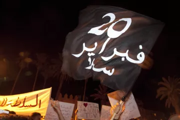 L'image montre une manifestation nocturne. À l'avant-plan, une personne tient un drapeau noir portant des inscriptions en blanc. Le drapeau semble être un symbole de protestation. En arrière-plan, on distingue des panneaux de revendications, également en arabe. L'atmosphère semble dynamique et engagée, illustrant un mouvement de contestation. La présence des palmiers suggère que l'événement se déroule dans un pays à climat chaud, probablement en Afrique du Nord.