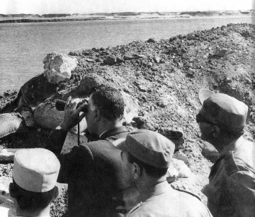 L'image montre un groupe d'hommes, dont l'un semble observer quelque chose au loin à l'aide de jumelles. Ils se tiennent près d'une rive d'eau, probablement à proximité d'un site de construction ou d'une digue, entourés de débris ou de terre. L'environnement semble suggérer une période historique, peut-être liée à des événements militaires ou politiques. Les hommes portent des uniformes ou des vêtements formels, ce qui indique qu'ils occupent probablement des positions officielles.
