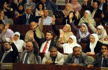 L'image montre un rassemblement dans un espace législatif, probablement au sein d'une assemblée ou d'un parlement. De nombreuses personnes sont présentes, avec une majorité levant la main, ce qui indique probablement un vote ou une approbation. Les participants semblent porter des tenues formelles, et certaines femmes portent le hijab. On observe une diversité de participants, tant en termes de genre que d'âge. L'environnement évoque une atmosphère de discussion et de prise de décision collective.