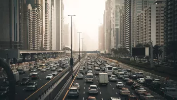 تظهر في الصورة مشهدًا حضريًا لمدينة مزدحمة، حيث تتواجد الكثير من السيارات المتوقفة في زحام مروري على عدة مسارات. تحيط بالمكان ناطحات السحاب العالية التي تدل على التطور العمراني في المنطقة. تبدو السماء مغشاة بضباب خفيف، مما يعطي انطباعًا عن أجواء المدينة في وقت متأخر من اليوم أو خلال الصباح الباكر. هناك مساحات من الأشجار على جوانب الطريق تضفي لمسة من الطبيعة وسط الحضر.