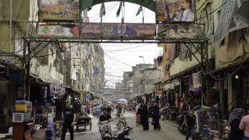 L'image montre une rue animée d'un quartier urbain. On peut y voir des bâtiments étroits et des fils électriques visibles. Des drapeaux et des affiches, probablement liés à un événement ou à une célébration, décorent l'entrée de la rue. Des personnes circulent, certaines à pied et d'autres à moto, ajoutant à l'activité de la scène. Le lieu semble être un marché ou un centre commercial, avec des étals vendant divers produits. L'atmosphère est vibrante et vivante.