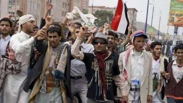 L'image montre un groupe de personnes en train de manifester dans une rue. Elles semblent exprimer des revendications, levant les bras et brandissant des drapeaux, notamment un drapeau rouge. Les manifestants portent des vêtements traditionnels yéménites, avec divers styles de vêtements. L'ambiance paraît énergique et engagée, comme une expression de protestation ou de soutien à une cause.