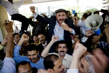 L'image montre une grande foule de personnes rassemblées, manifestant avec enthousiasme. Au centre, un homme souriant est porté sur les épaules d'un autre, levant le poing en signe de victoire. Les autres participants expriment également leur excitation, certains levant les bras et criant. La scène semble vibrante et dynamique, indiquant un moment de célébration ou de rassemblement collectif. Des panneaux et un mégaphone sont également visibles, suggérant une forme de discours ou de protestation.