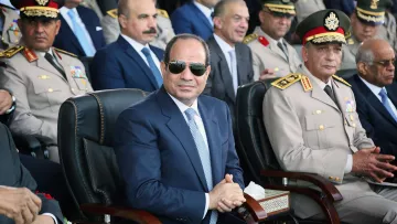 L'image montre un groupe de personnes lors d'un événement officiel. Au premier plan, un homme portant des lunettes de soleil et habillé d'un costume sombre est assis sur une chaise, semblant attentif. Il y a d'autres hommes en uniforme militaire et en tenue formelle derrière lui, qui semblent également être présents pour l'événement. L'ambiance est solennelle et il semble s'agir d'une cérémonie ou d'une célébration officielle.