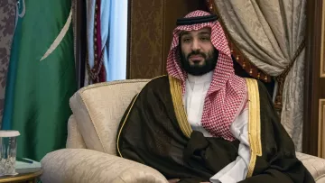L'image montre un homme assis sur un fauteuil, portant une tenue traditionnelle saoudienne, incluant une thobe blanche et un manteau noir avec des détails dorés. Il a une écharpe rouge et blanche, typique de la culture saoudienne, et il est entouré d'un décor riche, incluant des rideaux et des éléments décoratifs. On peut également apercevoir un drapeau saoudien en arrière-plan. L'expression de l'homme semble calme et posée.