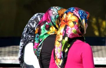 L'image montre le dos de trois femmes portant des foulards colorés. Chaque foulard est orné de motifs fleuris et vibrants, offrant un contraste frappant avec leurs vêtements. Les cheveux des femmes sont couverts, laissant entrevoir seulement leurs silhouettes. L'ambiance semble vivante et pleine de couleurs, soulignant la richesse culturelle des accessoires.