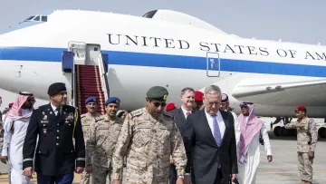 L'image montre un groupe de personnes déambulant sur un tarmac d'aéroport près d'un gros avion de couleur blanche et bleue, marqué "UNITED STATES OF AMERICA". Parmi eux, on peut voir des militaires en uniforme, des responsables en costume, ainsi que d'autres individus en tenue traditionnelle. Les hommes affichent des expressions sérieuses et semblent en discussion. L'appareil en arrière-plan suggère une visite officielle ou diplomatique.