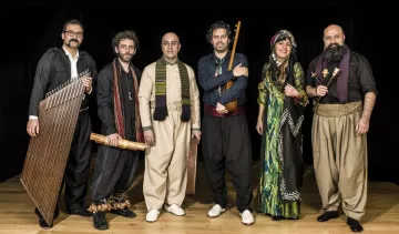 L'image montre un groupe de six musiciens posant ensemble sur scène. Ils sont vêtus de vêtements traditionnels, ce qui évoque une ambiance culturelle riche. Chaque membre tient un instrument différent : on peut voir un instrument à cordes, des percussions et d'autres instruments typiques. Leur expression est joyeuse et conviviale, et l'éclairage met en valeur leurs costumes. L'arrière-plan est sombre, ce qui fait ressortir leurs tenues colorées et détaillées. Cela donne une impression de performance musicale authentique et festive.