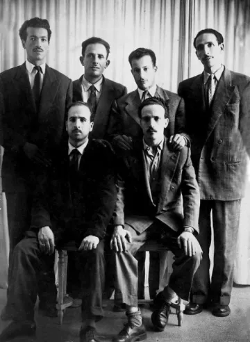 L'image montre un groupe de six hommes, habillés en costumes, posant ensemble. Ils sont disposés sur deux niveaux : trois debout à l'arrière et trois assis à l'avant. Tous les hommes affichent une expression sérieuse et se tiennent près les uns des autres, ce qui donne une impression d'unité. L'arrière-plan est neutre, avec des rideaux qui ajoutent une texture à la scène. La photo semble être en noir et blanc, ce qui lui confère un aspect vintage.