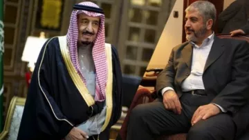 تظهر الصورة شخصين يجلسان في بيئة رسمية. الشخص على اليسار يرتدي لباساً تقليدياً يتضمن عقالاً وعباءة، مما يشير إلى هويته الثقافية. الشخص على اليمين يرتدي بدلة رسمية. يبدو أنهما يتبادلان الحديث في سياق دبلوماسي أو رسمي. الخلفية تضم تفاصيل معمارية تشير إلى مكان مهم.