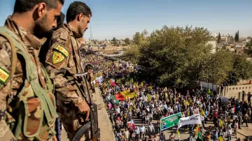 L'image montre une scène de manifestation en plein air, où une grande foule est rassemblée, tenant des drapeaux et des pancartes. Au premier plan, deux soldats armés, portant des uniformes camouflés, surveillent la foule depuis un point élevé. On peut voir des arbres et des bâtiments en arrière-plan, et le ciel est dégagé. L'atmosphère semble chargée d'émotion, avec des participants visiblement engagés.
