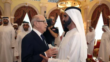 L'image montre deux hommes en discussion, probablement dans un cadre formel et décoré, avec des éléments architecturaux typiques d'un palais. L'un des hommes porte un costume sombre, tandis que l'autre est vêtu d'une robe traditionnelle blanche. En arrière-plan, on aperçoit d'autres personnes, également en tenue traditionnelle, qui semblent observer la conversation. L'ambiance semble conviviale et respectueuse.
