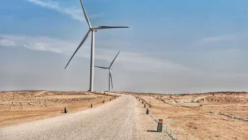 The image depicts a wide, unpaved road extending through a dry, barren landscape. On either side of the road, there are several large wind turbines standing tall against a clear blue sky. The terrain appears arid, with sparse vegetation and a sandy or rocky surface. The road is lined with small posts or markers, adding a structured element to the otherwise natural setting. The overall atmosphere conveys a sense of open space and renewable energy in a desert-like environment.