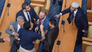 L'image montre une scène tendue dans une assemblée ou un parlement. Plusieurs hommes discutent vivement, certains pointant du doigt et affichant des expressions agitées. Il y a une mise en scène de désaccord, avec des gestes éloquents et une proximité physique entre les participants. Le décor est simple, avec des sièges en bois et un tapis bleu au sol, suggérant un environnement formel.