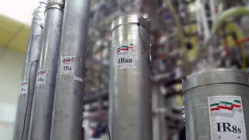 La imagen muestra varios cilindros metálicos dispuestos en un laboratorio o instalación industrial. Cada cilindro tiene una etiqueta que indica "IR8" o "IR8b", probablemente refiriéndose a modelos de centrifugas. En las etiquetas se puede ver una representación de la bandera de Irán. El fondo está desenfocado, pero se pueden observar más estructuras relacionadas con el equipo técnico, sugiriendo un ambiente relacionado con la investigación o la producción.