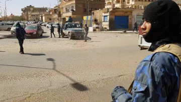 L'immagine mostra una scena di una zona urbana, presumibilmente in un contesto di sicurezza elevata. In primo piano, un uomo indossa un berretto e abbigliamento camuffato, mentre tiene un'arma. Sullo sfondo si possono vedere altri uomini in uniformi simili che si muovono in un'area in cui ci sono veicoli parcheggiati. L'architettura circostante appare tipica di una città, con edifici vari e attività commerciali. La luce del giorno illumina la scena, creando ombre ben definite.