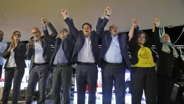 L'image montre un groupe de personnes sur une scène, levant les bras en l'air dans un geste de célébration ou de triomphe. Ils semblent être réunis pour un événement politique, probablement après l'annonce de résultats électoraux. Les participants portent des vêtements formels, et l'ambiance paraît festive et unie. En arrière-plan, on peut voir des écrans avec des informations, ce qui suggère que c'est une occasion importante.