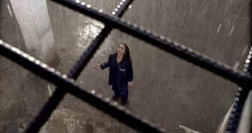 L'image montre une scène dramatique d'une femme debout dans un espace clos, probablement une cellule. Elle porte une tenue de prison et semble être sous une forte pluie qui tombe du plafond. La lumière est tamisée, créant une ambiance sombre et mélancolique. La femme, les bras ouverts et la tête inclinée vers le ciel, exprime une pose de désespoir ou de lâcher-prise, tandis que des barreaux en fer témoignent de son confinement. Le sol semble humide et dégradé, ce qui renforce l'atmosphère oppressante de l'environnement.