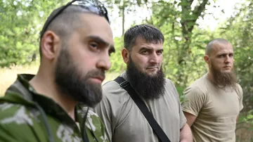 L'image montre trois hommes se tenant côte à côte dans une forêt. Tous trois portent des vêtements utilitaires, et chacun d'eux a une barbe. Leurs expressions sont sérieuses, et l'éclairage naturel met en valeur leur présence dans ce cadre boisé. On peut observer des arbres en arrière-plan, ce qui crée une ambiance naturelle.