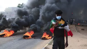 L'image montre un jeune homme marchant sur une route pendant une manifestation. Il porte un masque et fait un signe de paix avec sa main. En arrière-plan, on peut voir des pneus enflammés et une épaisse colonne de fumée noire s'élevant dans l'air. La scène dégage une impression de tension et de protestation, et il y a un groupe de personnes visibles derrière lui.