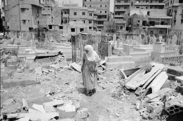 L'image montre une femme vêtue d'un long habit, marchant au milieu d'un cimetière. Le sol est désordonné, avec des débris éparpillés, ce qui suggère qu'il y a eu des destructions récentes dans la zone. En arrière-plan, on peut voir des bâtiments, probablement endommagés, qui entourent le cimetière. L'atmosphère paraît lourde, évoquant des sentiments de tristesse et de désolation.