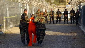 L'image montre un environnement militaire, probablement un centre de détention. On y voit des soldats en uniforme camouflé, certains tenant un individu vêtu d'une combinaison orange, qui semble être escorté. À l'arrière-plan, d'autres soldats sont alignés, surveillant la scène. Le tout se déroule dans un espace clôturé, avec des barbelés visibles, ce qui accentue l'atmosphère de sécurité et de contrôle. La lumière est naturelle, suggérant un moment en journée.