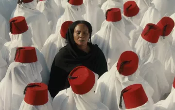 L'image montre un groupe de personnes vêtues de longues robes blanches, portant des fez rouges. Au centre, une femme en noir se tient les yeux fermés, contrastant avec l'uniformité des autres. Cette composition crée une atmosphère de méditation ou de réflexion, avec un fort accent sur la spiritualité et la communauté. Les couleurs et les vêtements évoquent une tradition culturelle riche.