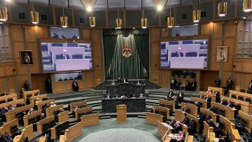 L'image montre l'intérieur d'une grande salle de réunion ou d'assemblée, probablement un parlement ou une assemblée législative. Au centre, une estrade est prévue pour les orateurs, et des écrans sont visibles sur les murs pour diffuser les interventions. On peut voir plusieurs membres assis sur des rangées en pente, organisés en sections. Les couleurs dominantes sont des tons de vert et de bois, créant une ambiance formelle. Des personnes sont également présentes debout, écoutant attentivement.