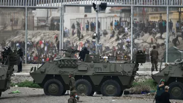 La imagen muestra un contexto de tensión, probablemente en una zona de conflicto o crisis. Se pueden ver vehículos militares blindados en la parte frontal, con soldados a bordo y personal militar en el entorno. Al fondo, hay una multitud de personas que parecen estar frente a una valla alta, indicando una separación entre diferentes grupos. El ambiente es de intensa actividad, posiblemente con indicios de enfrentamientos o una situación de disturbio. La escena evoca un fuerte sentido de vigilancia y seguridad militar.