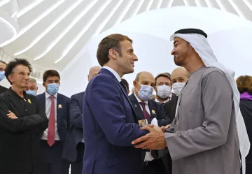 The image depicts two men engaged in a handshake, likely in a formal setting. One individual is dressed in a suit, while the other is wearing traditional attire, possibly indicating a cultural exchange. Surrounding them are several other people, some of whom are wearing face masks. The background features a modern architectural design, contributing to the contemporary atmosphere of the scene.