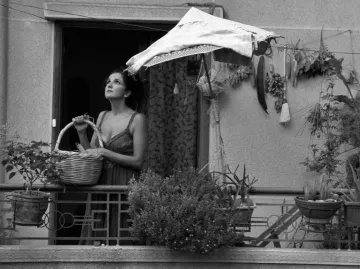 L'image montre une femme se tenant sur un balcon, tenant un panier dans ses bras. Elle semble contemplative, regardant au loin. Le balcon est décoré de plusieurs plantes et herbes suspendues. Un parasol en toile protège en partie la femme du soleil. L'image est en noir et blanc, ce qui lui donne une atmosphère nostalgique et mélancolique.
