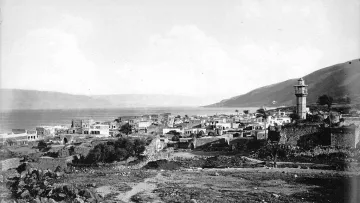The image depicts a historical view of a coastal town near a body of water, likely a lake or sea, with a prominent lighthouse tower standing tall in the background. The landscape features a mix of stone buildings and ruins, suggesting a blend of architecture that may reflect the area's history. Surrounding the town are hills or mountains, adding depth to the scene. The overall mood is serene and nostalgic, capturing a moment from the past with a monochromatic or sepia tone.