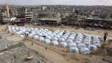 L'immagine mostra un'ampia area con numerosi tende bianche disposte in file ordinate, che rappresentano un campo per sfollati. Sullo sfondo si possono vedere edifici distrutti e in rovina, segno di conflitti o disastri. Il cielo è nuvoloso, contribuendo a un'atmosfera desolante. La scena evidenzia il contrasto tra le tende temporanee, che offrono riparo, e l'ambiente urbano devastato circostante.
