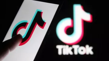 این تصویر یک گوشی هوشمند را نشان می‌دهد که لوگوی اپلیکیشن TikTok روی صفحه آن قابل مشاهده است. انگشت یک دست به سمت لوگو اشاره می‌کند. در پس‌زمینه، لوگوی TikTok به صورت بزرگ‌تر و با رنگ‌های آبی و قرمز دیده می‌شود. طراحی این لوگو به‌صورت مدرن و جذاب است و نشان‌دهنده‌ی ماهیت سرگرم‌کننده اپلیکیشن می‌باشد.