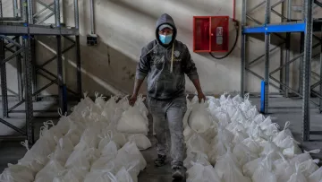 L'immagine mostra un uomo che cammina in un ambiente di magazzino. Indossa una felpa con cappuccio e una mascherina e tiene in mano due sacchetti bianchi. Intorno a lui ci sono numerosi sacchetti bianchi disposti ordinatamente sul pavimento. Le pareti del magazzino sono spoglie e ci sono scaffali metallici sullo sfondo. L'atmosfera sembra lavorativa e organizzata.