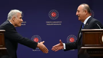 L'image montre deux hommes se préparant à se serrer la main dans un cadre officiel. Ils se tiennent devant un fond avec des symboles de la République de Turquie et des inscriptions officielles. L'un des hommes porte un costume sombre et semble sourire, tandis que l'autre a également un costume et une expression sérieuse. L'atmosphère dégage une impression de diplomatie et de protocole.